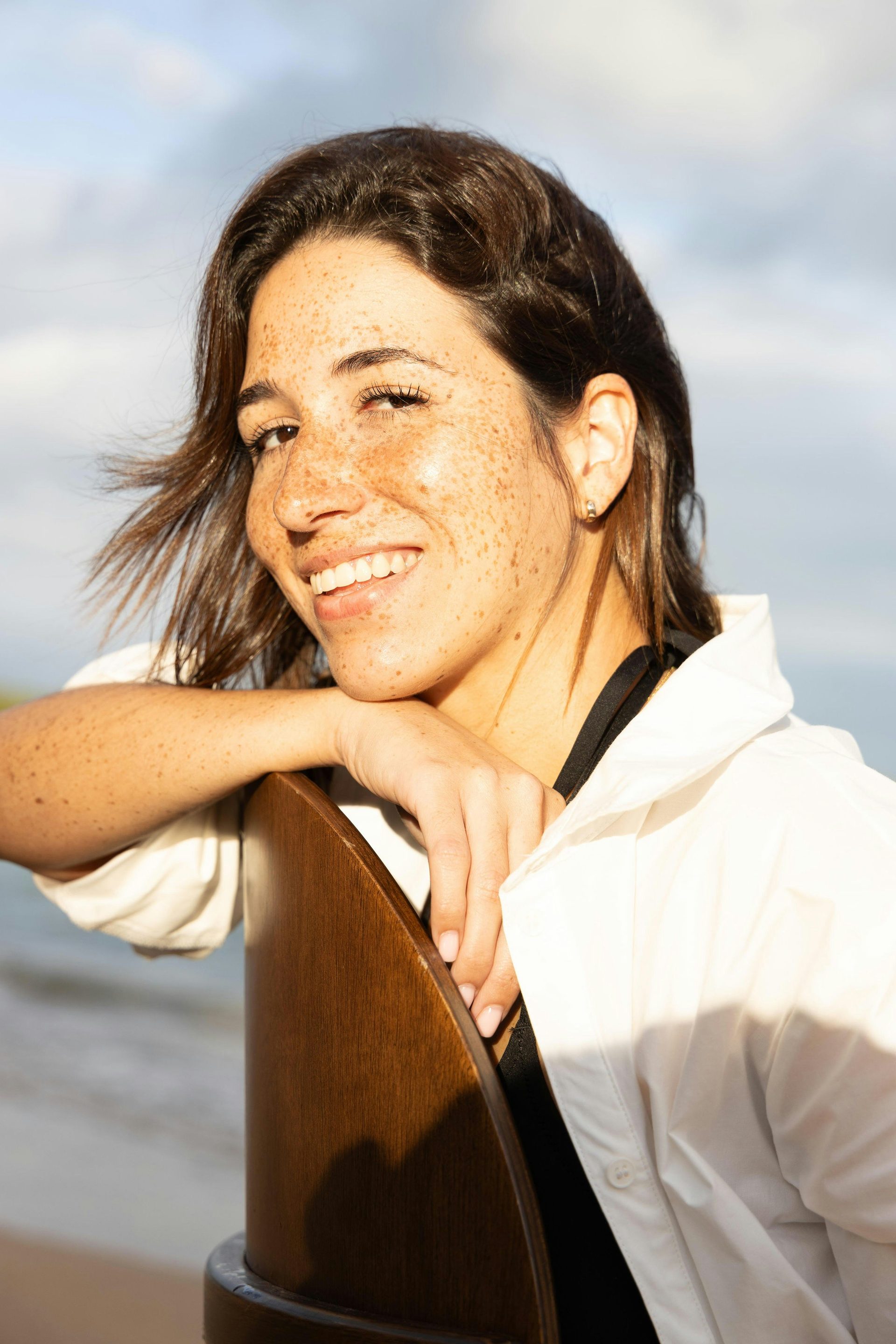 A freckled woman leaning on a chair by the sea, smiling