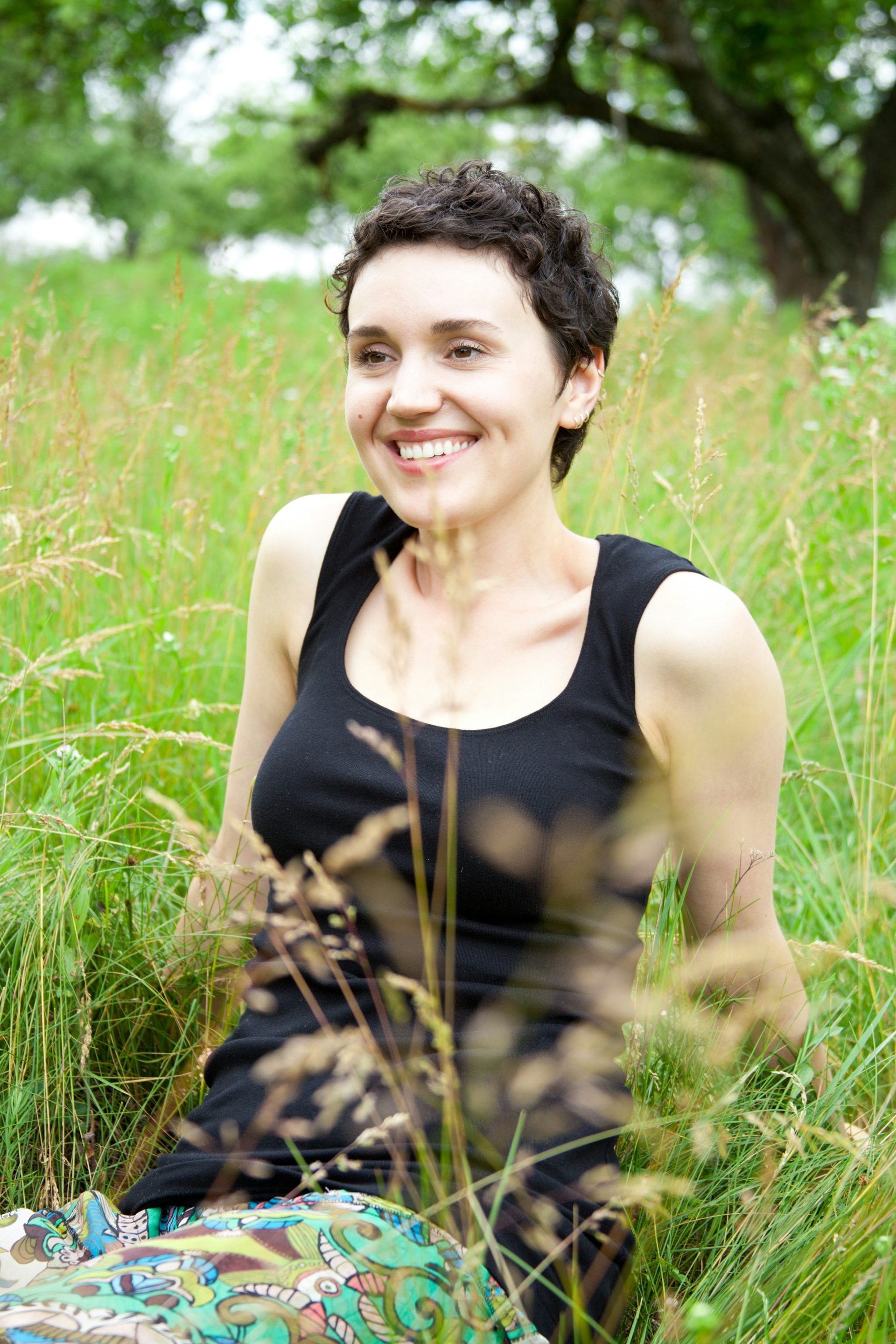 A woman with dark curls sitting in tall grass, smiling
