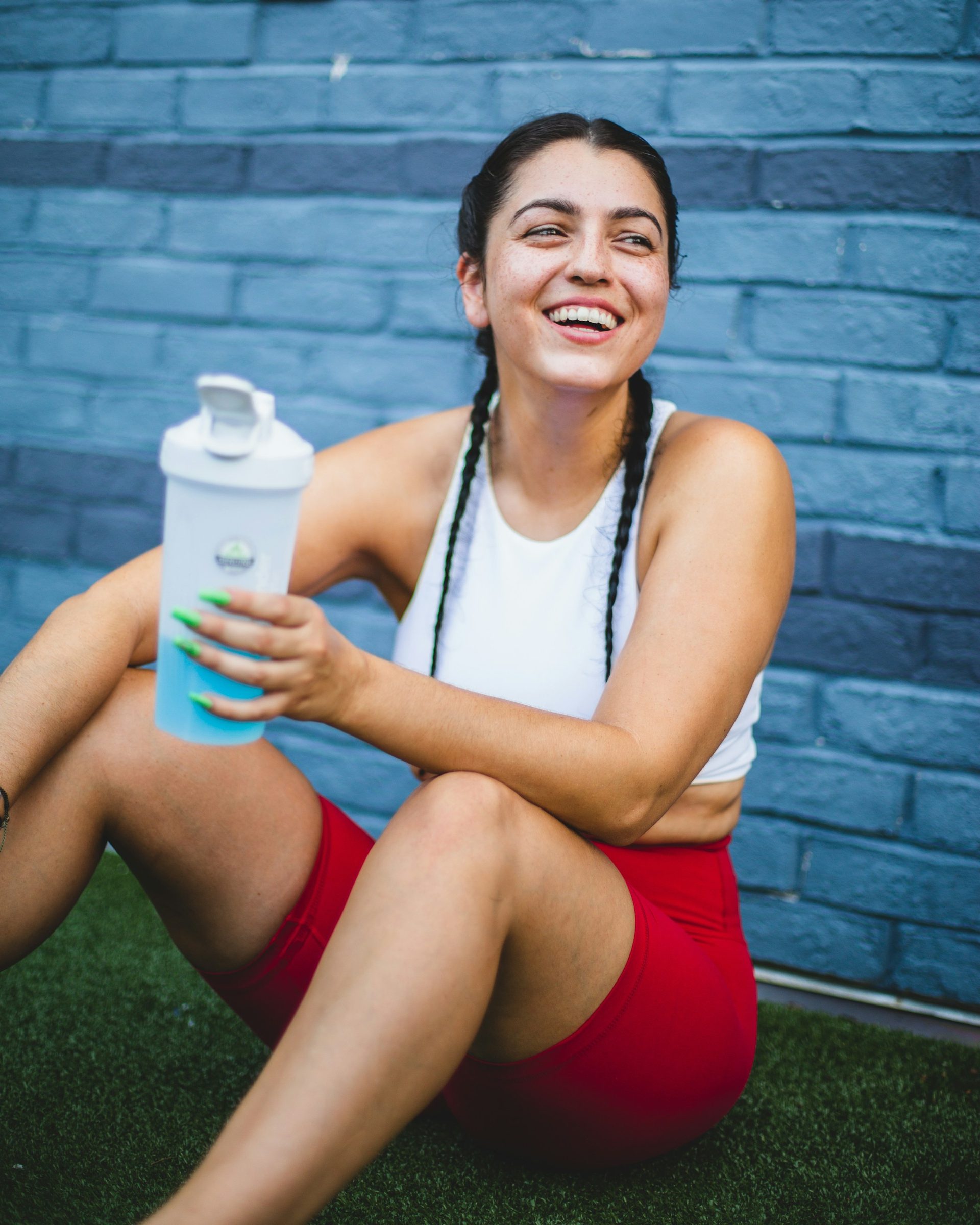 A woman in workout clothes sitting on turf, smiling, drink bottle in hand