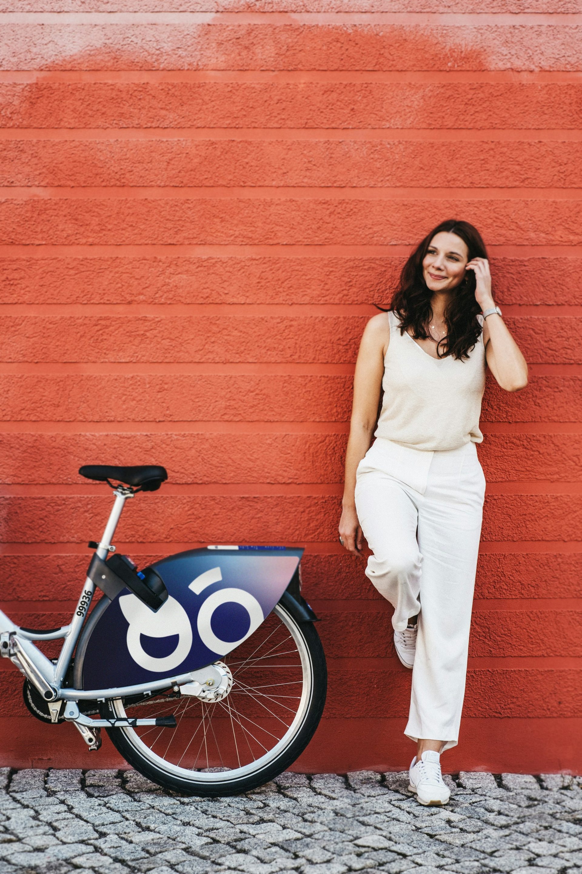 A woman in white leaning against a red wall beside a bike