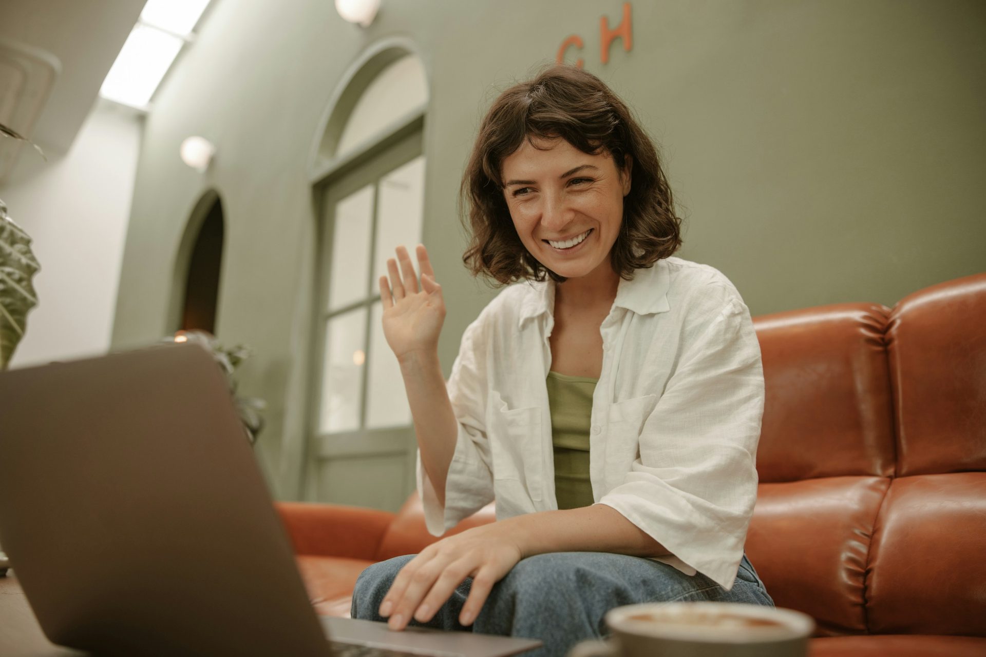 A woman on a sofa waving at a laptop during a video call