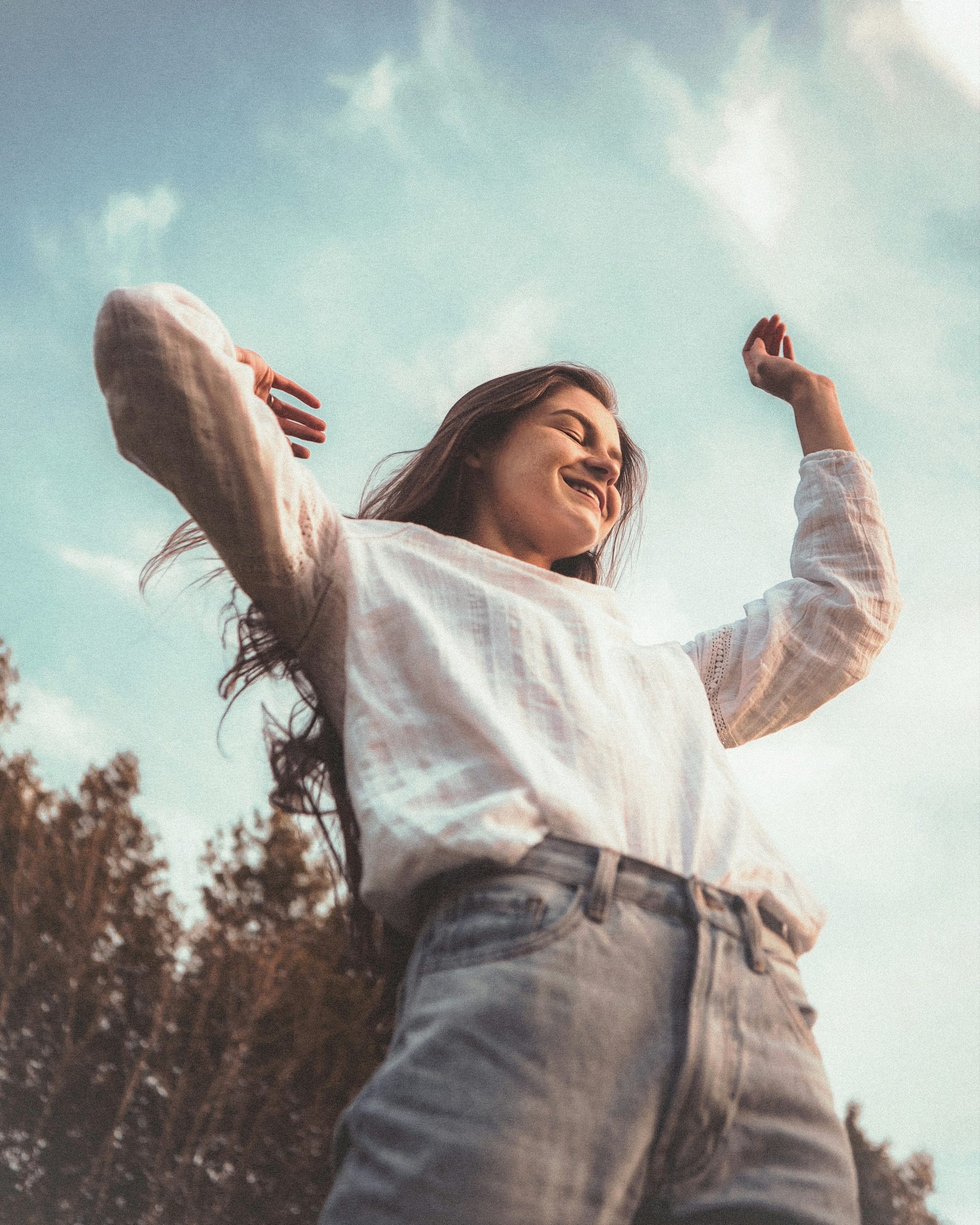 A woman in a white shirt, arms raised and smiling, under an open sky